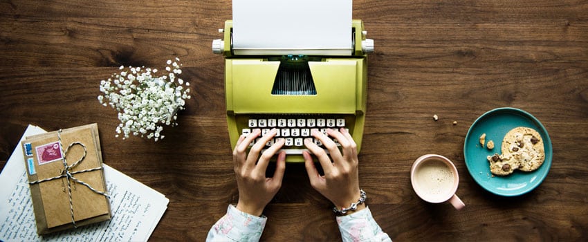 Woman's hands typing in the typewriter; Left side of the typewriter are envelopes tied with a knot, paper with writings, and flowers; On the other side are a cup of coffee and a plate with cookies