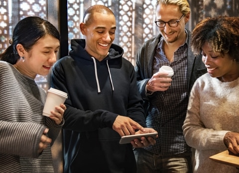Group of friends chatting together, the white girl on the right holding a cup, the bald man pointing on his phone, the second man staring at the phone and holding a cup, and the black woman looking at the phone, having an elegant background.