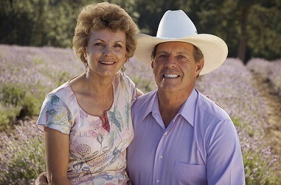 Old Couple sitting together. Old man have a white cowboy hat and blue polo at right, old woman in dress at left. In barren land