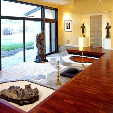 Meditation room. Front has glass doors and wall, right side has concrete walls, with frame, 2 statue and at the center of 2 statue is a glass squared wall. wooden floor, below the wooden floor is tile floor with carpet, 2 candles and between 2 candles is a square pillow with round pillow at the top, a figurine at the left side, statue near to the glass door