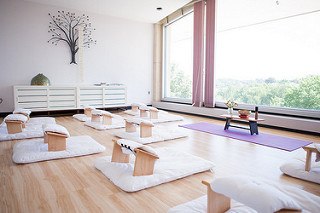 A room with wooden floor, glass wall at the back, concrete white wall at the right side with tree decor hanged, white shelves. White mats with small wooden chairs with foam on the floor. At the center is a purple mat with small wooden table with vases and bowl on the top.