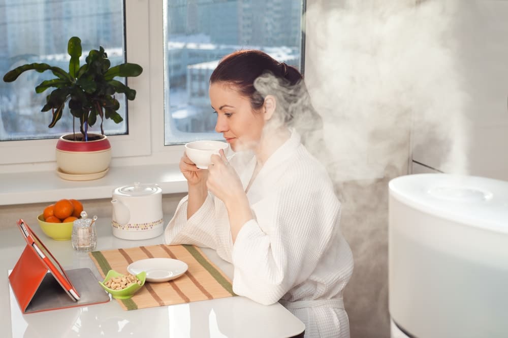 Woman wearing a white bathrobe, drinking tea reading tablet at humidifier while looking on her tablet