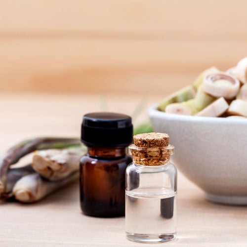 Chopped lemongrass in a bowl, lemongrass plant below the bowl and 2 small bottles of oil in light brown background