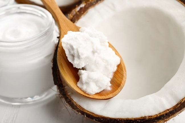 A spoonful of processed coconut oil placed above a husked coconut with jar full of cream on the corner