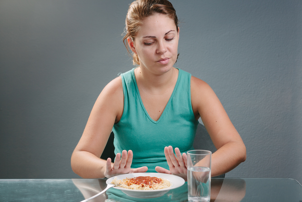 A middle-aged woman with a brown hair, wearing a blue green sleeveless shirt, sitting on the chair in front of the table having no appetite in front of the meal. Concept of no appetite woman with a gray background.