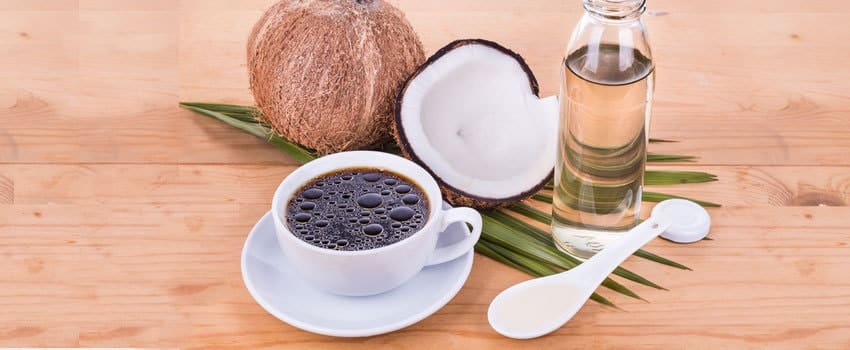 A coffee, coconut shell with its flesh intact, a bottle of coconut oil, and a spoon- all arranged pleasantly on a wooden table