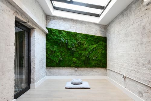 Concrete meditation room. Glass door on the left side, at the front is a large green plants painting, glass ceiling, tile floor with square pillow and a round pillow at the top.