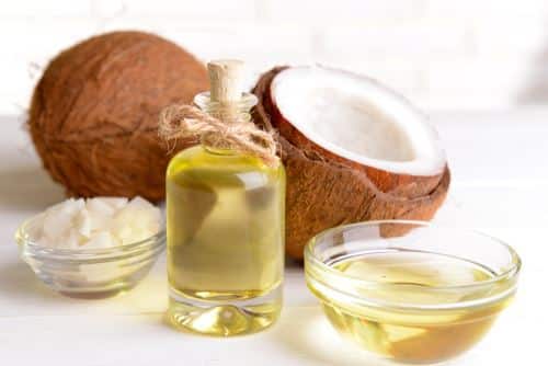 A coconut oil inside a clear bottle with a cover and a ribbon. A bowl of coconut chunk. A bowl of coconut oil. A whole coconut beside the half coconut fruit with shell with a white background.