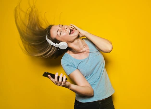 A woman with blonde hair wearing a blue blouse and black jeans, wearing a headphone, and holding a black cellphone in yellow background. A woman singing.