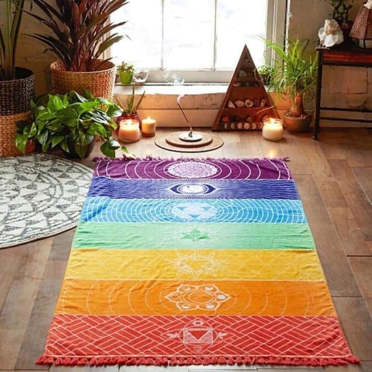 Chakra Tapestry Colorful Rainbow Design on the Floor. Meditation room. Wooden floor with rainbow carpet, circle carpet, knitted pot with flowers, a glass window at the top, scented sticks, candles, flower vase with flower, a chair