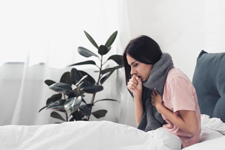 Woman with gray scarf on her neck sitting down on the bed, coughing while touching her chest inside the room