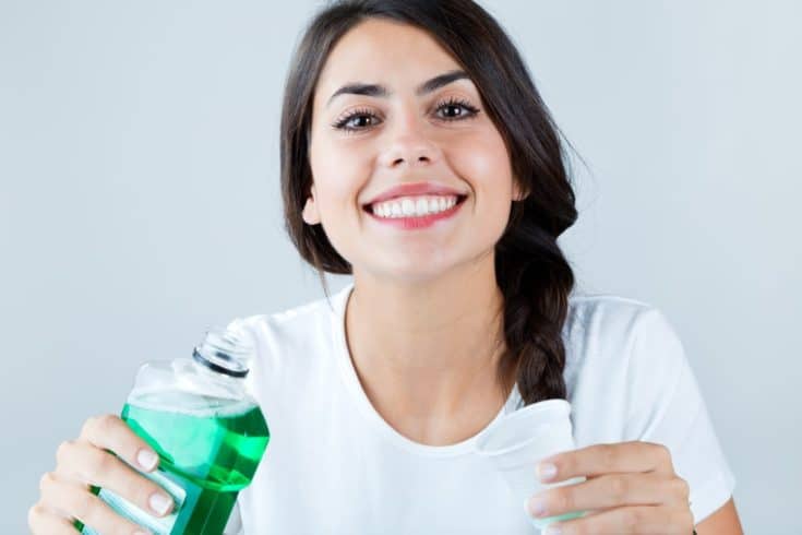 Portrait of a beautiful girl wearing white t-shirt, holding a mouthwash and a white cup. Isolated on white.