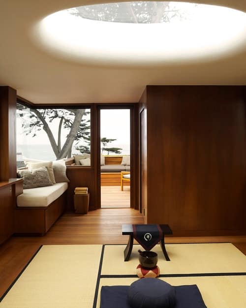 Japanese meditation room. Wooden room with glass window, beige carpet on the floor with small wooden chair with cloth, a bowl placed in round stone, square pillow with round pillow at the top. Near the door is a wooden chair with foam at the top and pillows. Outside is a tree and a terrace