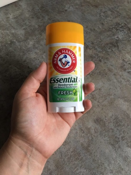 A person's hand holding the Arm & Hammer Essentials Deodorant in a grey concrete background