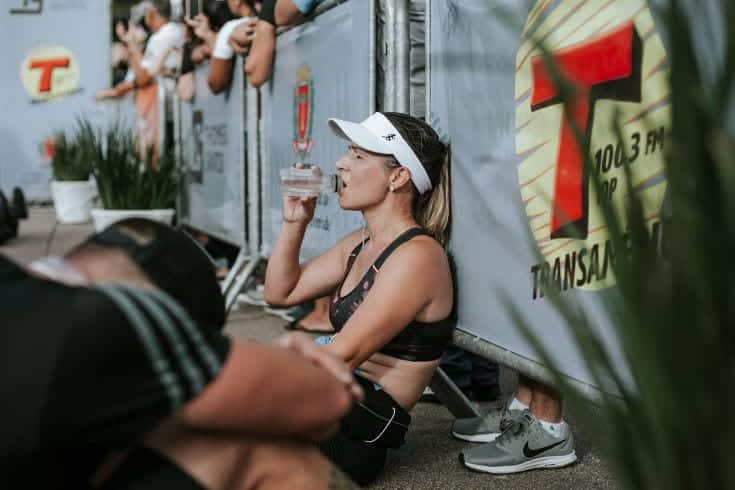 athlete woman sitting down on the ground while drinking water with blurry people background