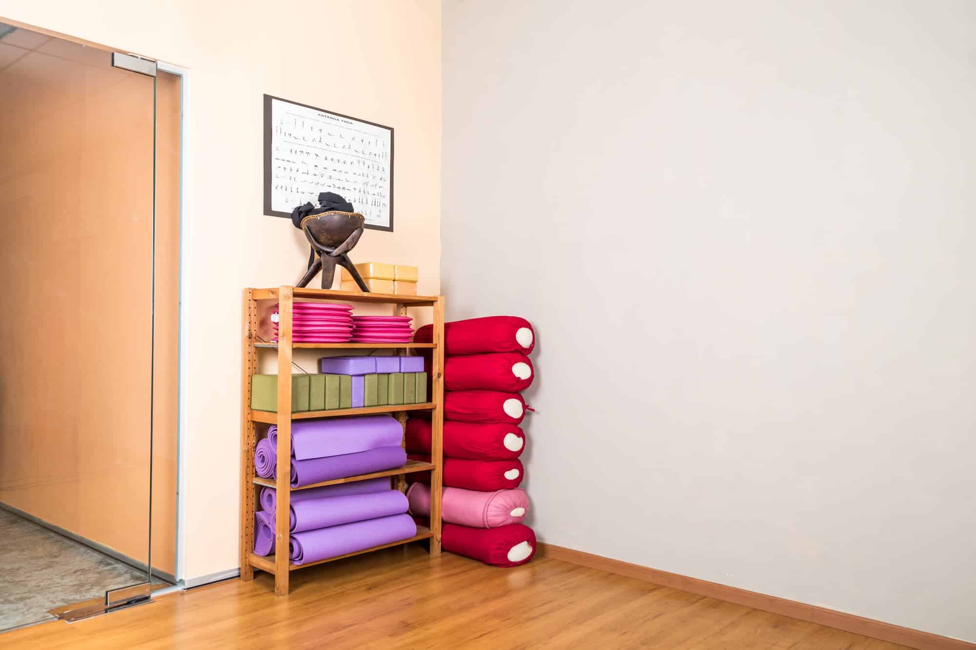 Yoga blocks, pillow, mats, pads, accessories stacked in empty yoga studio with wooden flooring